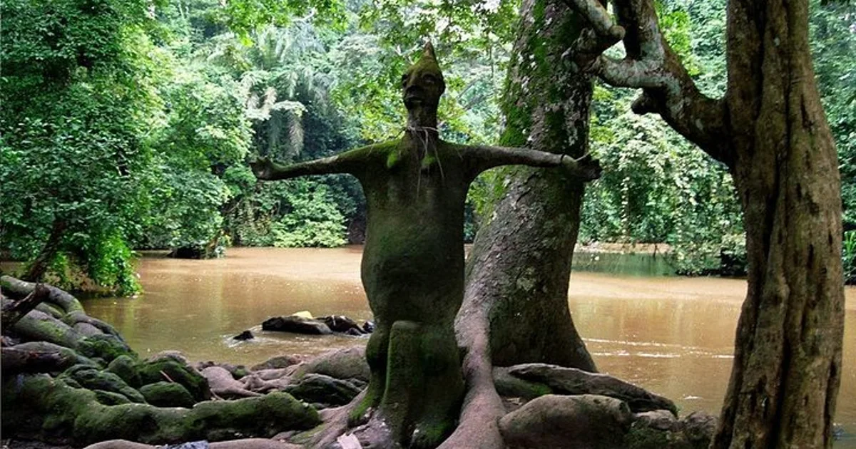 A large sacred sculpture beneath forest trees beside the Osun River at Osun-Osogbo Sacred Grove.