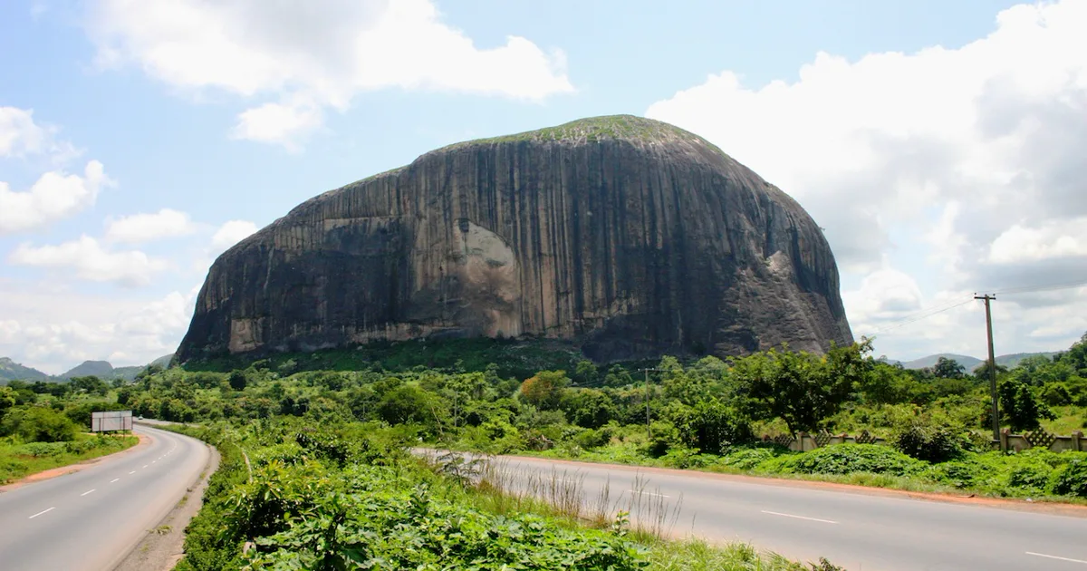 Zuma Rock rising beside the Abuja–Kaduna road beneath a bright blue sky.